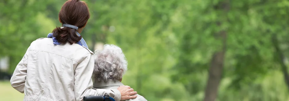 A woman with her arm around an elderly person, both facing a lush, green park. The scene conveys warmth and support amidst a tranquil, natural backdrop.